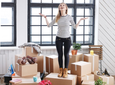 Woman standing on cardboard boxes in a bright room, gesturing with hands, surrounded by moving supplies, reflecting Octopus Moving Company's services in Belmont, MA.